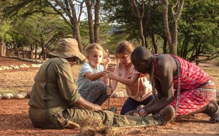Two children work together with a camp guide and local Maasai warrior to create a fire.