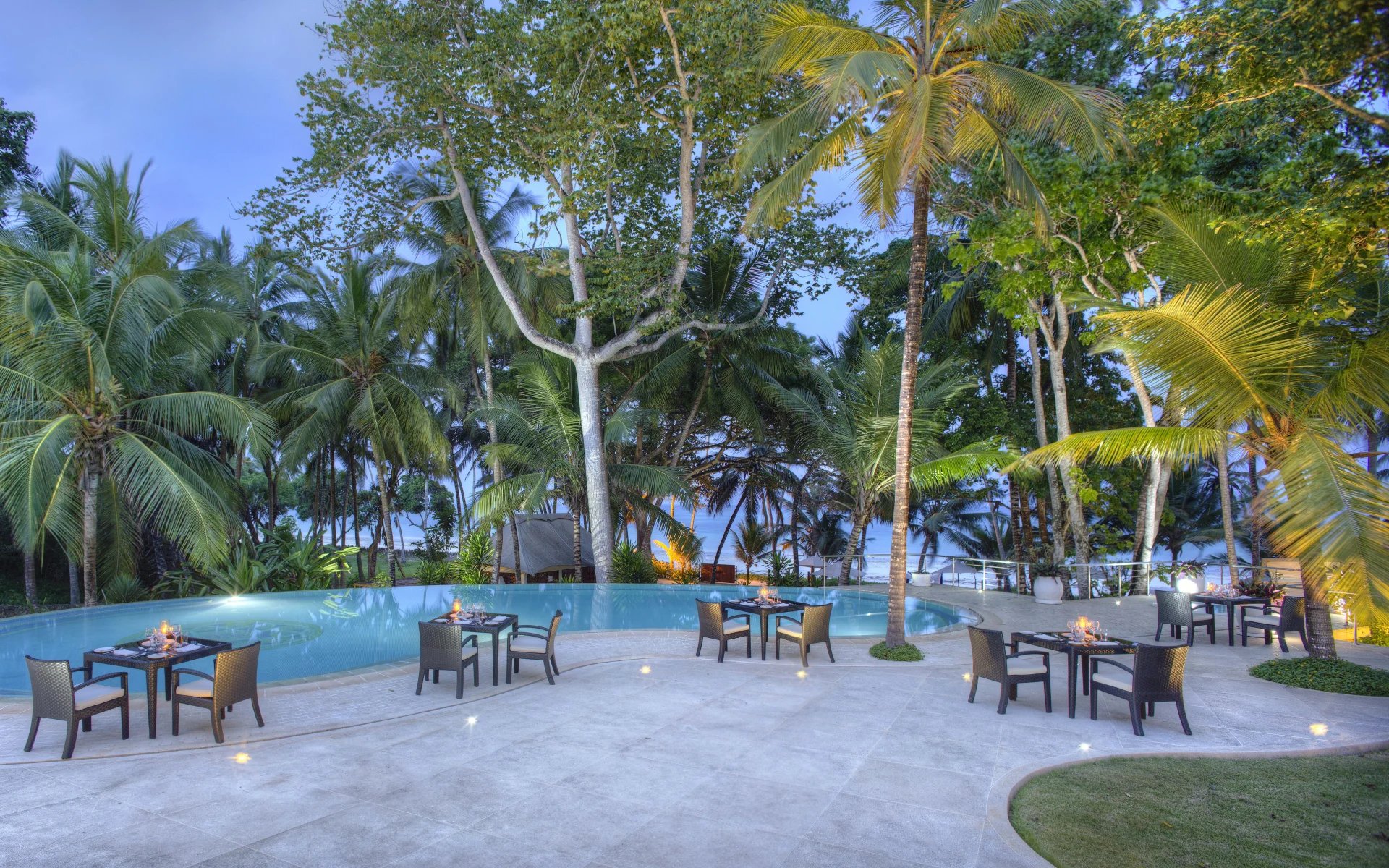 Chairs and tables are dotted around the Almanara's outdoor pool, encased by tall, tropical trees and plants.
