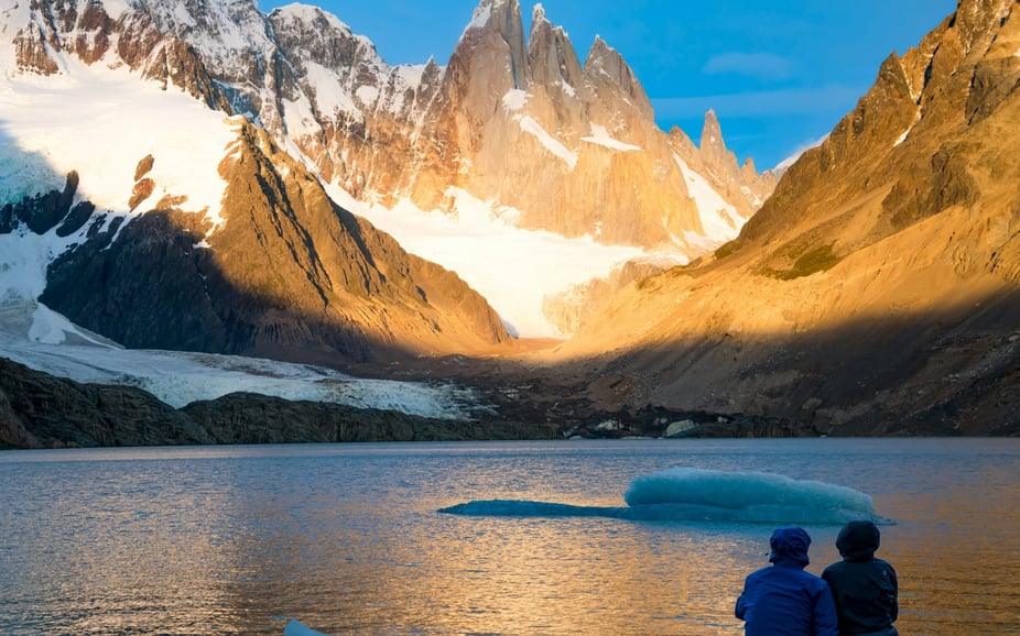 couple sat beside lake with small ice berg in the middle surrounded by moutains
