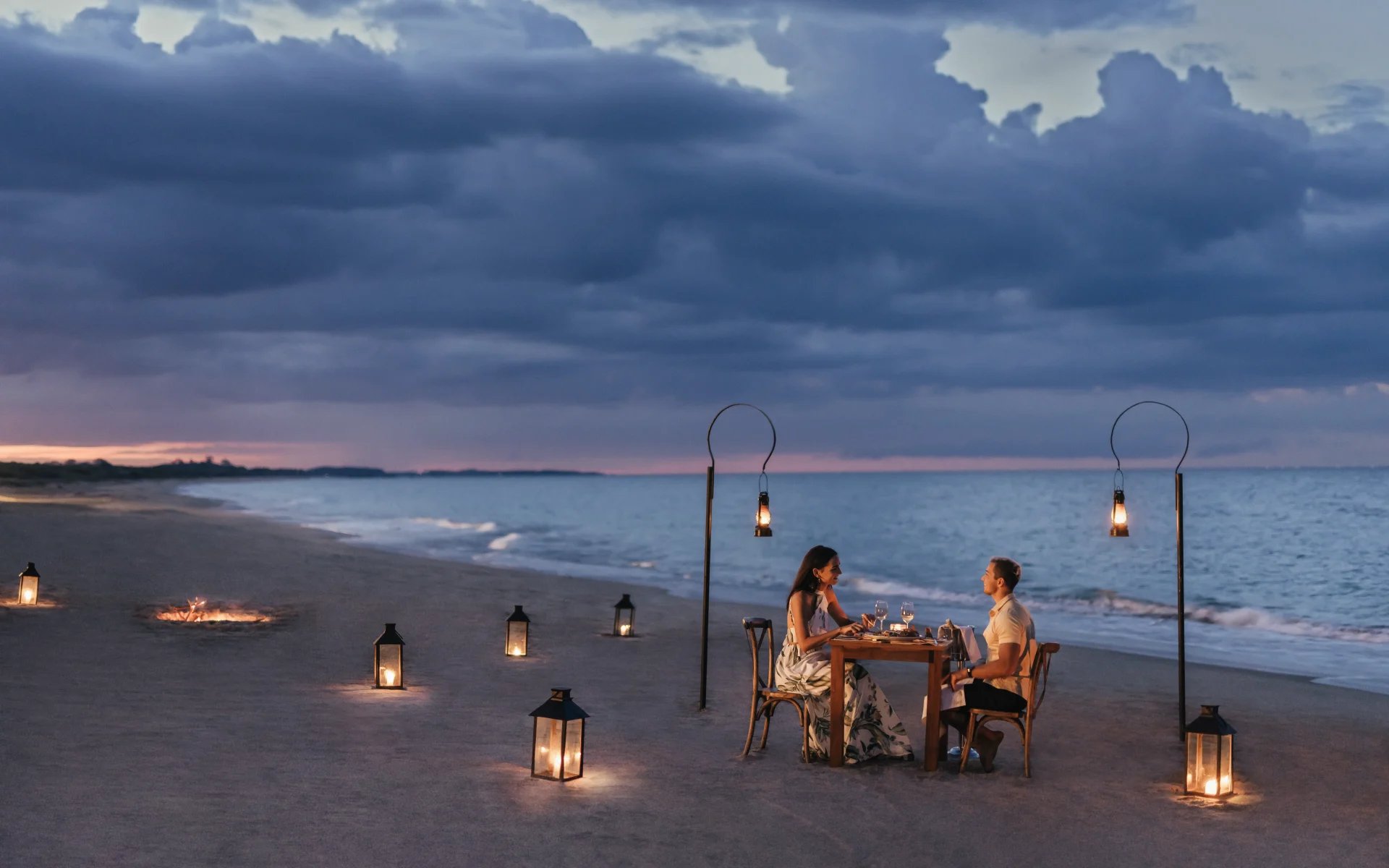 A couple is sitting at a table on the beach at nighttime, surrounded by lanternlights. 