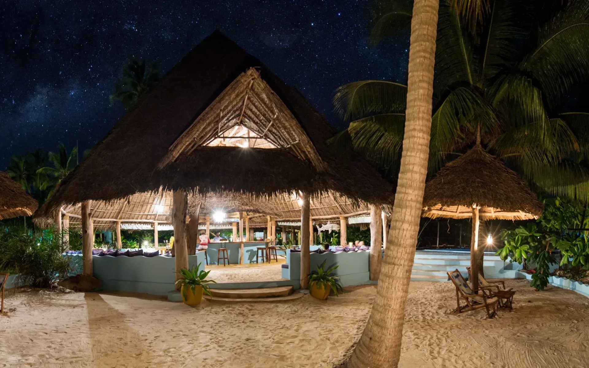 The thatched-roof beach bar at Butiama Beach Lodge is lit up by soft lighting during the evening. It is surrounded by tall palms.