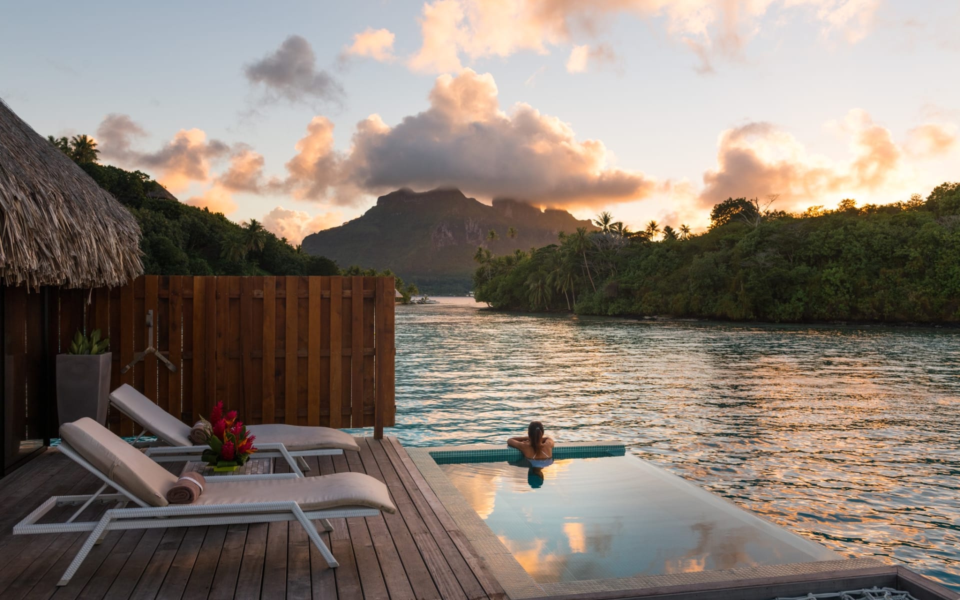A woman is sitting in an infinity pool, watching the sun set over a massive mountain. 
