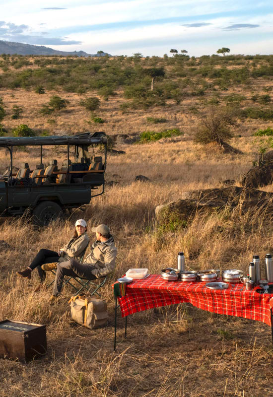A couple relax on camping chairs beside a picnic table with flasks during a daytime game drive.