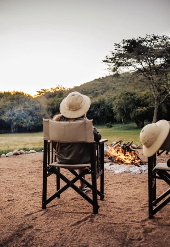 Two people relax on chairs ahead of a campfire during the early evening, wearing safari hats.