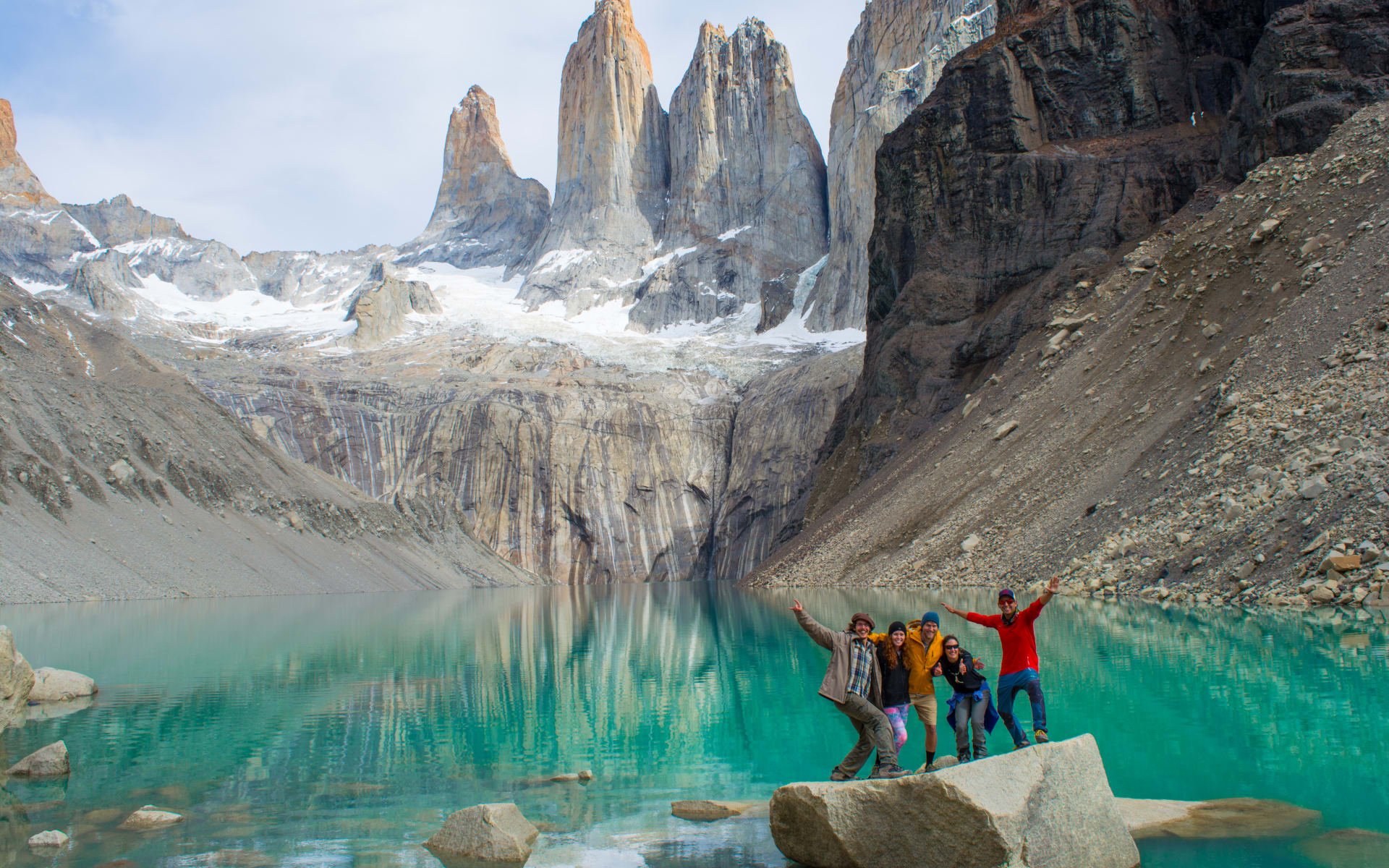 A group of people are standing in front of a turquoise lake with snow-capped mountains in the background. 
