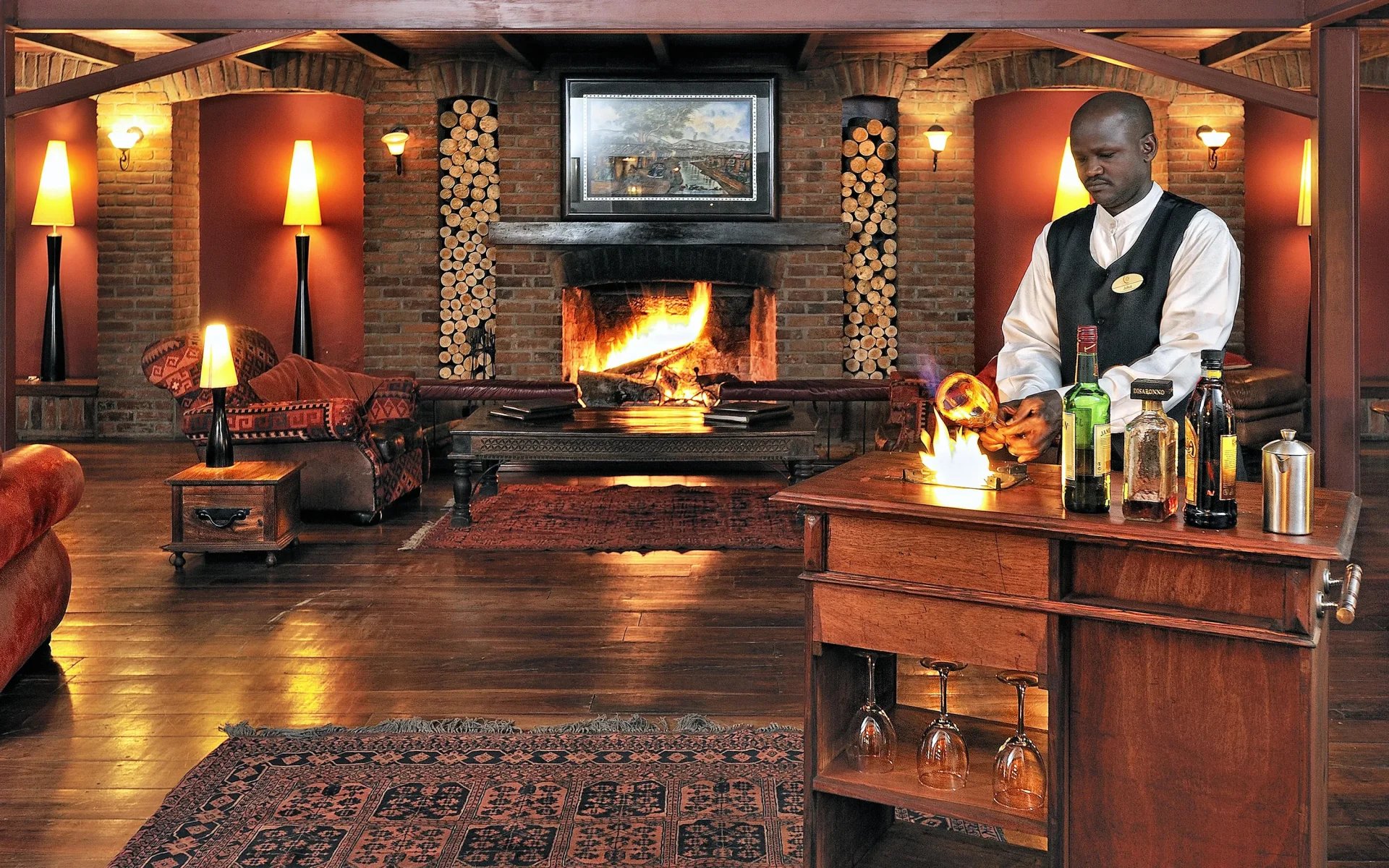 A waiter carefully prepares cocktails in a wood-panelled room which is centered by a logfire and a tapestry rug.