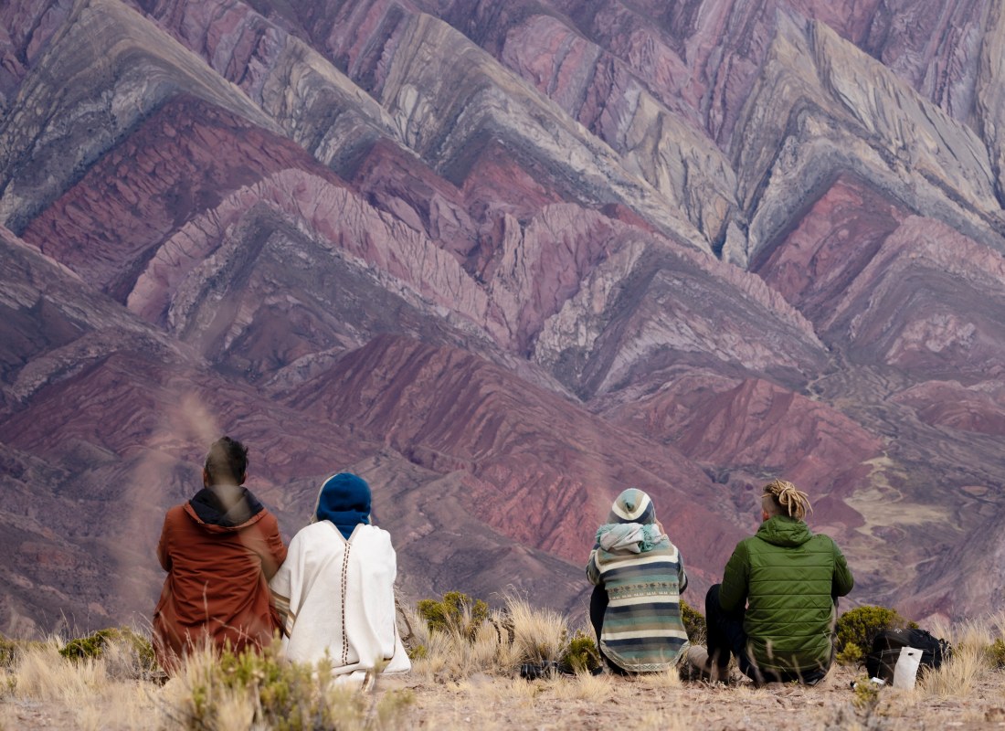 A group of four are sitting on the dry grass overlooking the multi-coloured mountains in Humahuaca.
