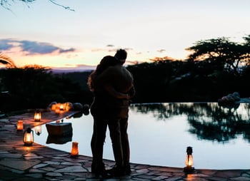 A couple cuddle on the poolside, surrounded by candlelit lamps ahead of a gentle sunset.