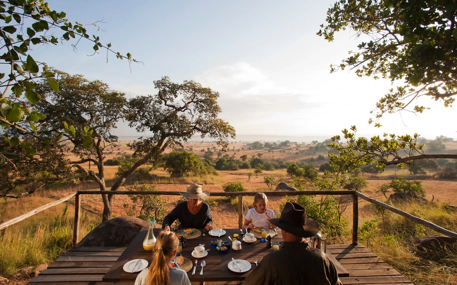 A family enjoy a delicious breakfast on the sun deck at Lamai Serengeti, ahead of a vast scene of grasslands and hillsides.