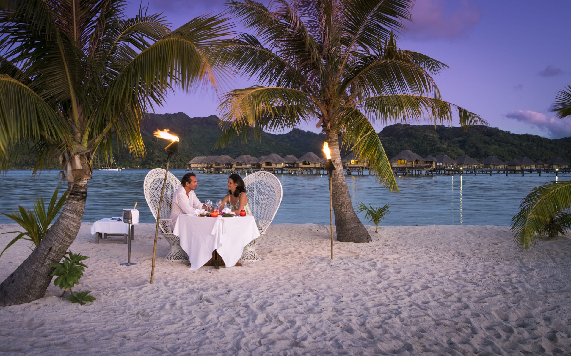 A couple are sitting at a table on the beach as the sun sets. 