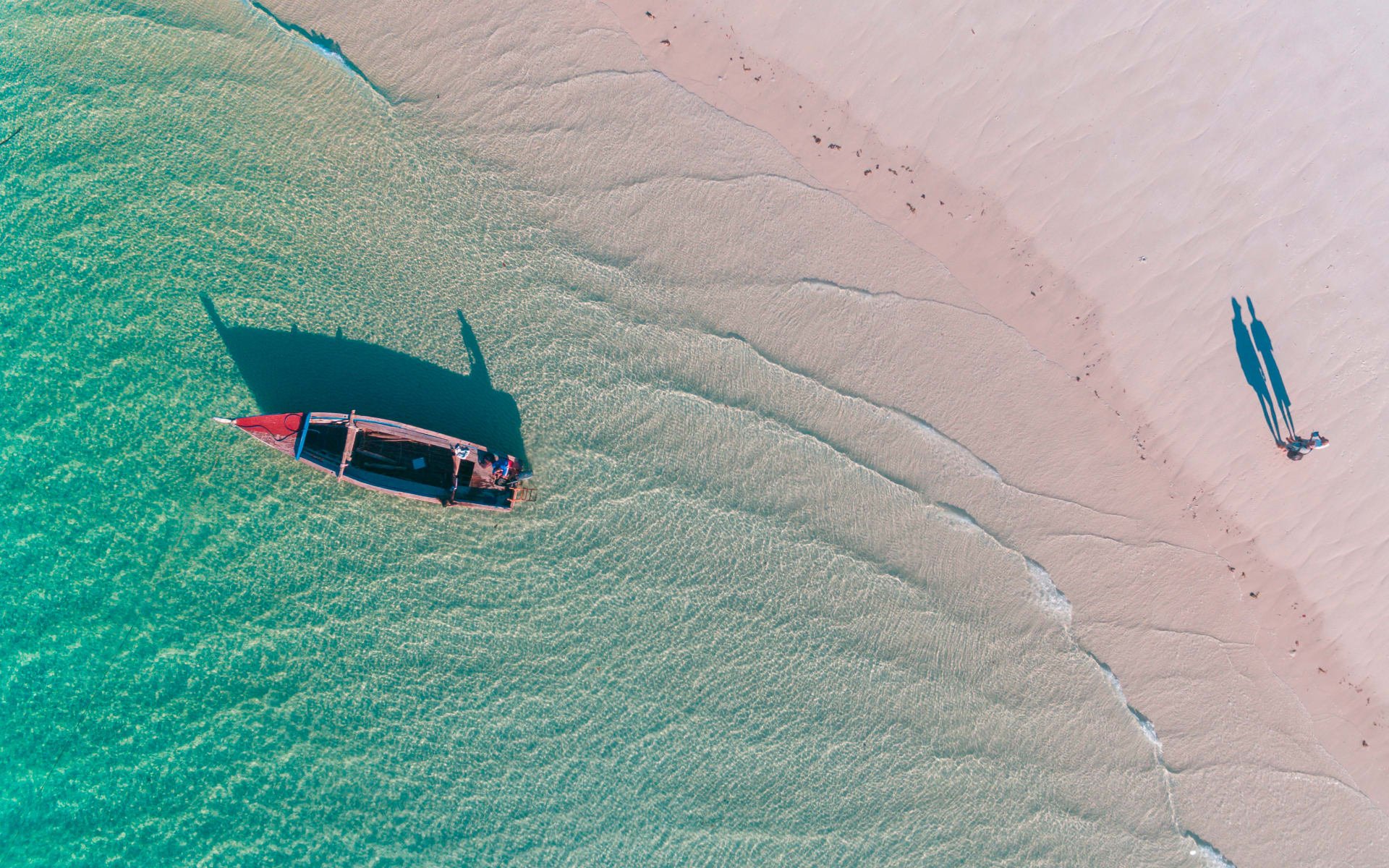 An aerial image of a couple walking along the sandy ocean shore as a small fishing boat floats by.