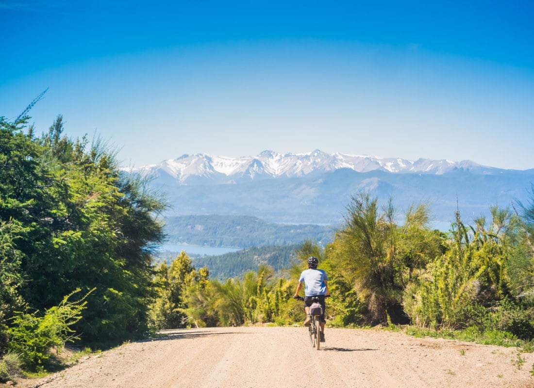 A man is cycling along a dirt road with Patagonia's snow-capped mountains in the distance.