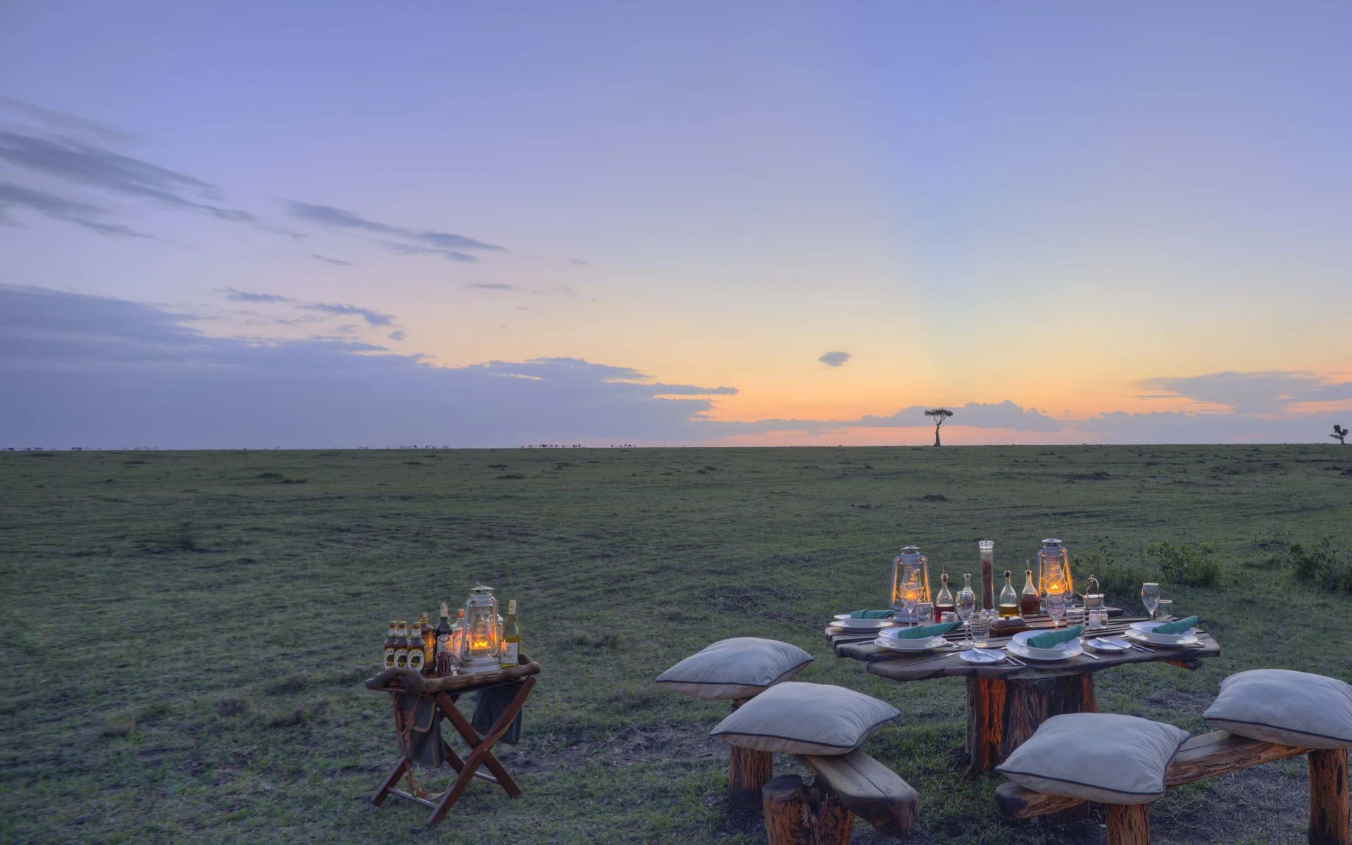 A private dining arrangement is set up ahead of an orange sunset on the Masai Mara plains.