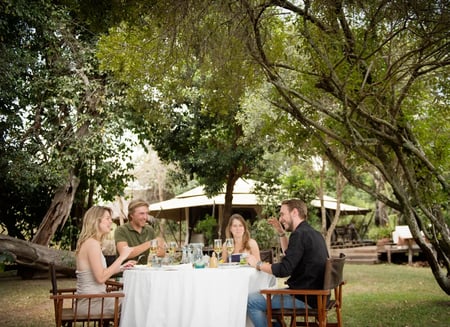A group of four friends sit in a tree-sheltered area enjoying wine and lunch as they laugh together.