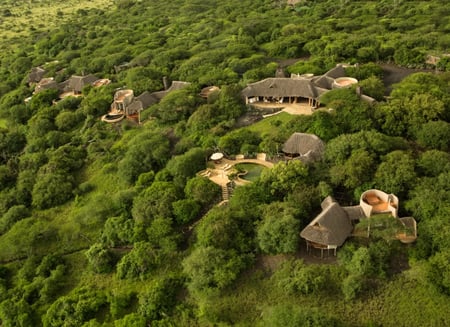 An aerial view of Ol Donyo Lodge shows the lodge's thatched-roof buildings and outdoor pool encased by thick foliage.