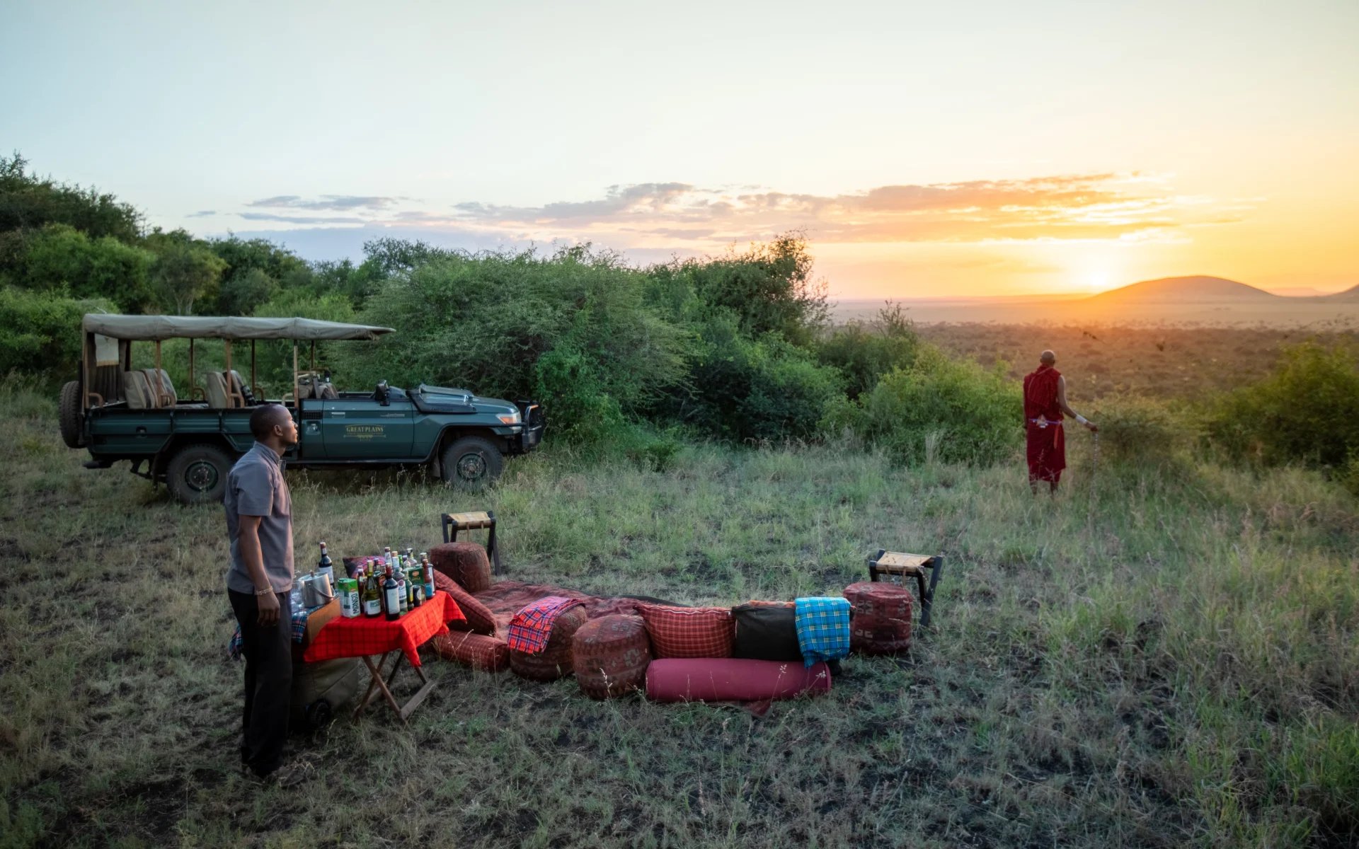 A picnic is set out on a hill on the reserve during sunset. A camp guide and local Maasai warrior admire the view.