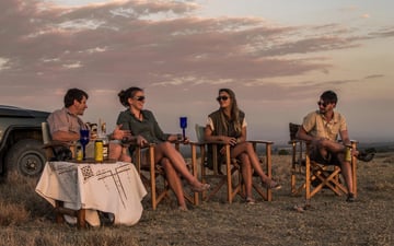 Four people are sitting in deck chairs next to a safari vehicle and a white-clothed table. 
