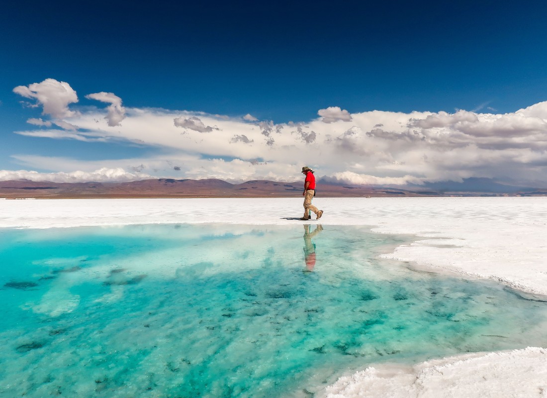 A man in a red coat is walking across Salinas Grande Salt Flats' white terrain, and he's reflected in a turquoise pool. 