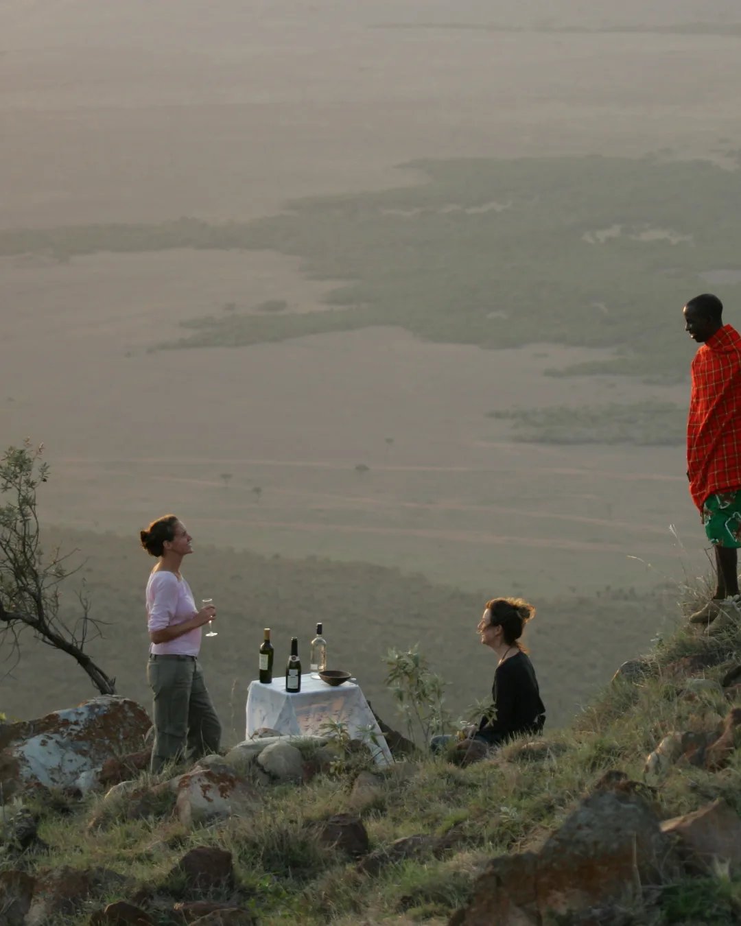 A tribal guide is standing on a rock with two guests who are having a picnic overlooking the plains. 