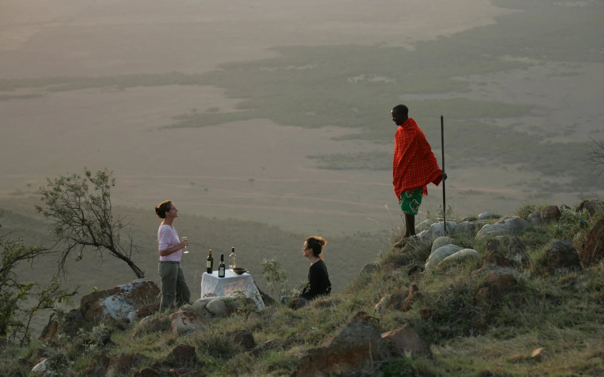 A local Maasai man speaks to a couple as they enjoy a private dining experience on a cliffside in the Masai Mara.