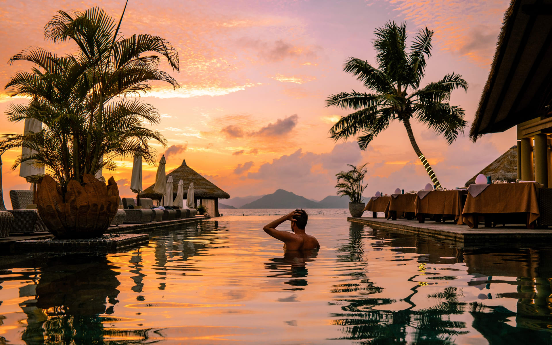 A man bathes in an outdoor infinity pool ahead of a pink sunset.