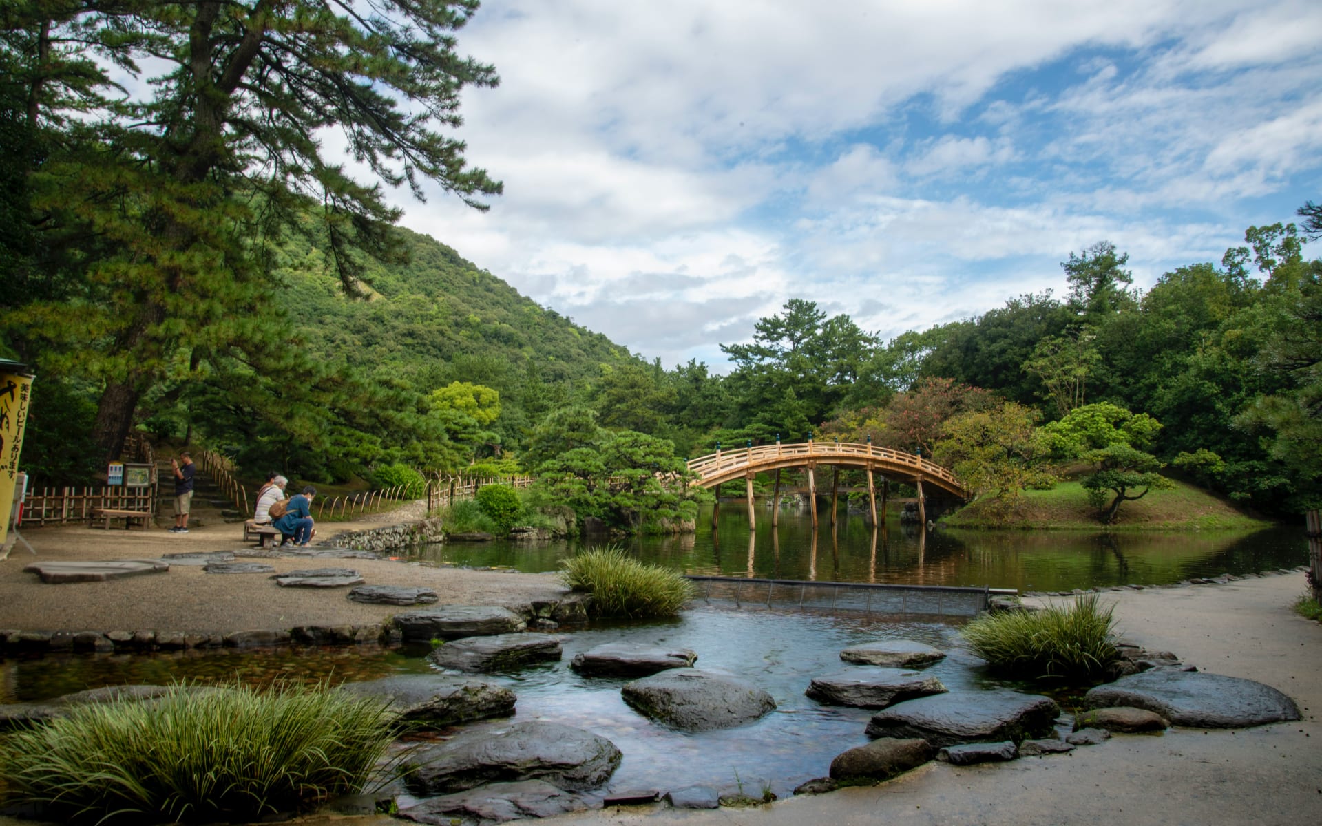 A couple are sitting on a wooden bench in Takamatsu park, overlooking a bridge and river.