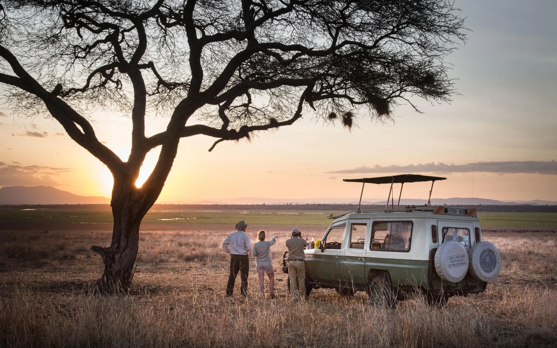 A couple and a tour guide stand beside their game vehicle ahead of a gentle sunrise, beside a large acacia tree in Tarangire National Park.
