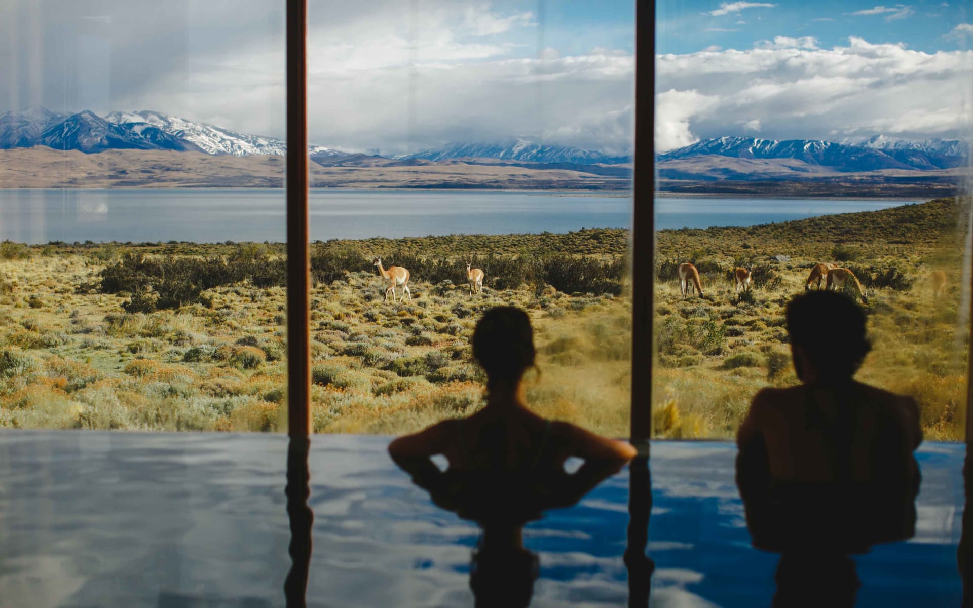 Two people are looking through a glass window from an indoor pool.