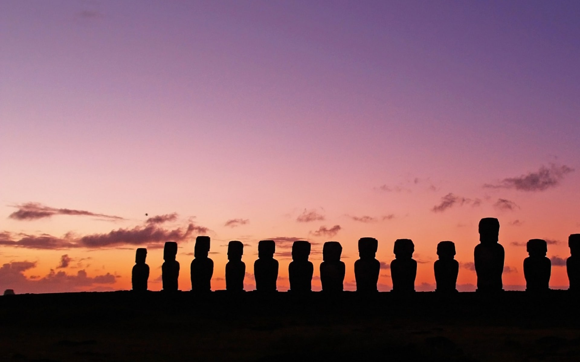 Moai statues in Easter Island stand in a row while the sun sets, cascading orange, yellow and purple hues across the sky.