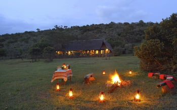 There is a fire pit with logs and lanterns surrounding it on the lawn of Saruni Basecamp Mara Lodge.