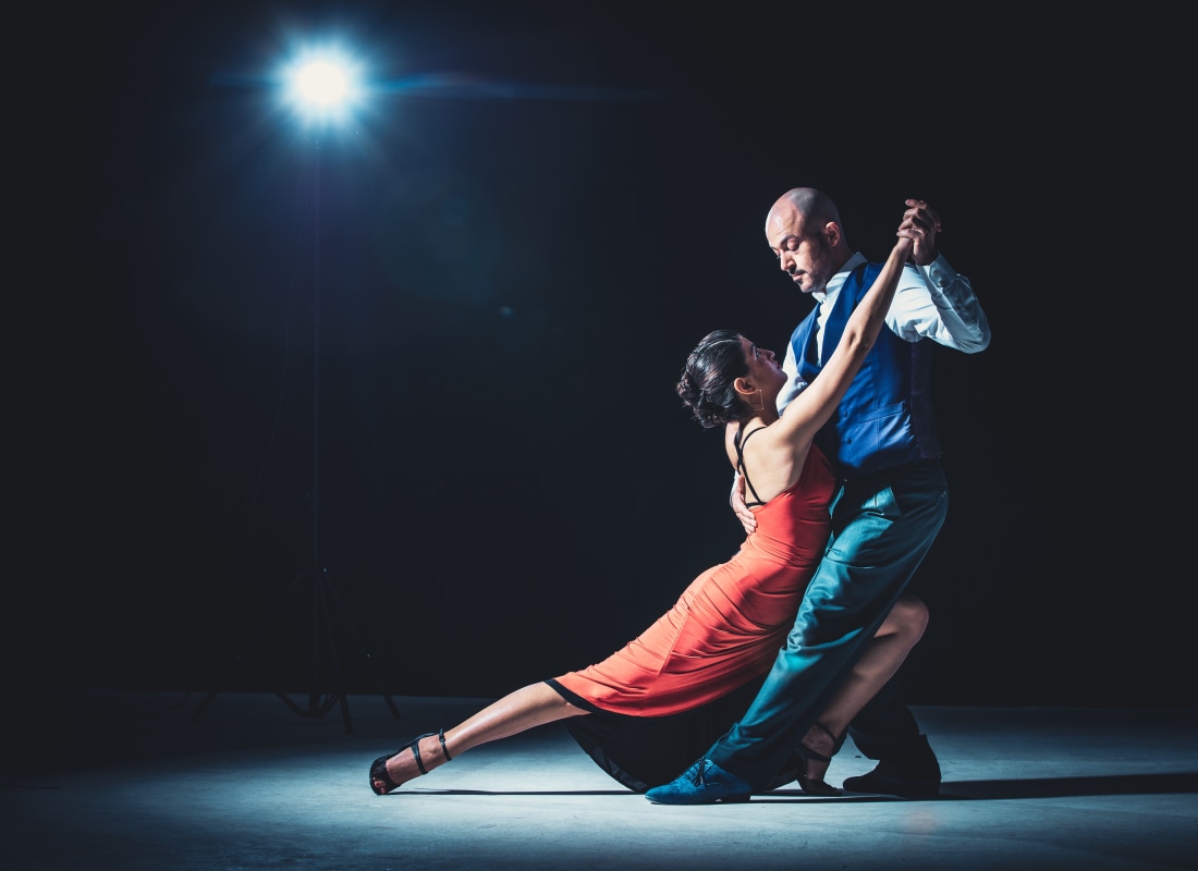 A man and woman in a red dress are dancing the tango in a black room and a spotlight on on them. 