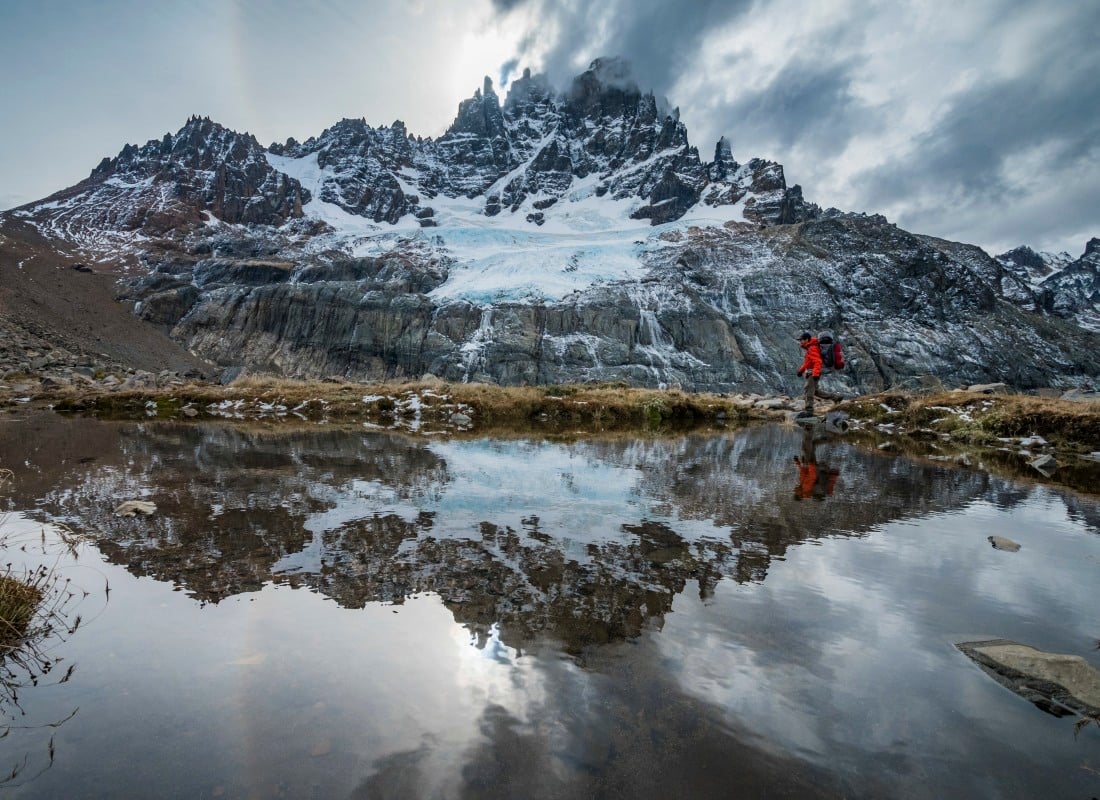 A man is hiking in the mountains, walking over a lake backed by snow. 