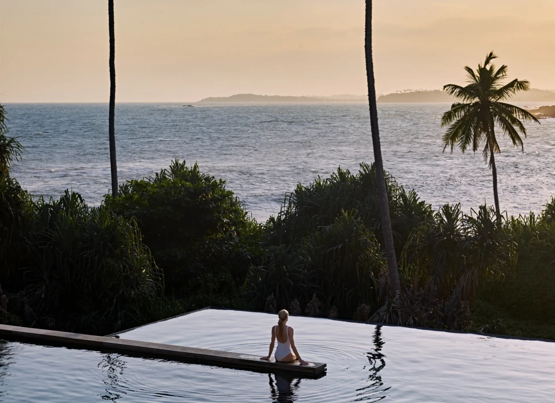 A woman is sitting in an infinity pool overlooking the ocean.