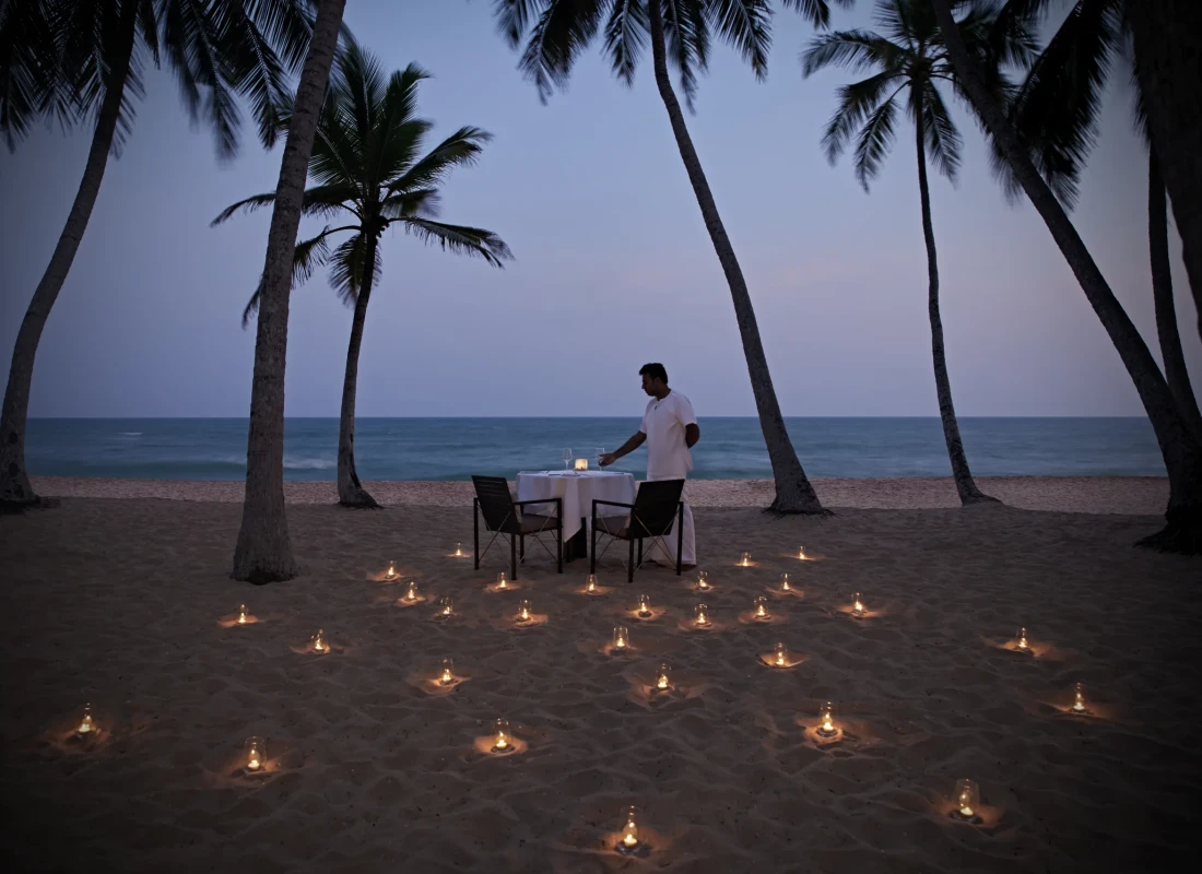 A man is lying at the table on the beach with candles and the ocean surrounding him. 