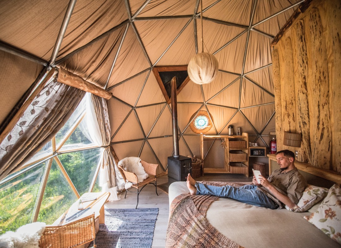 A man sits on a cosy bed in a dome with a large window overlooking the Patagonian countryside. 