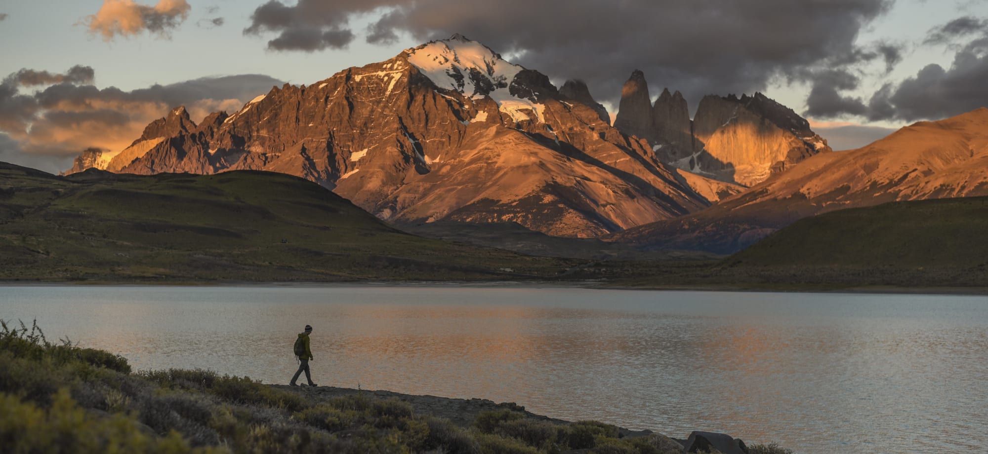 A man is walking in front of a lake with mountains in the background glowing orange from the sunset. 