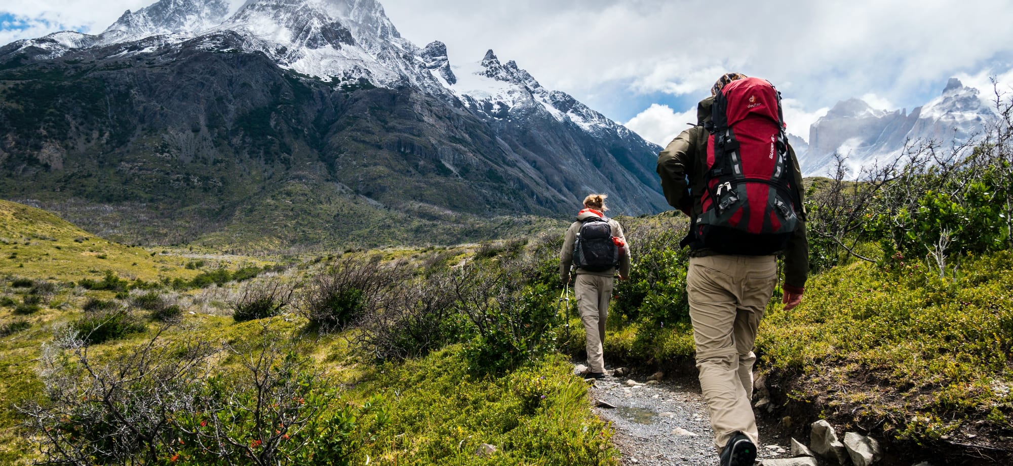 A couple are hiking in Patagonia, surrounded by snow-capped mountains. 