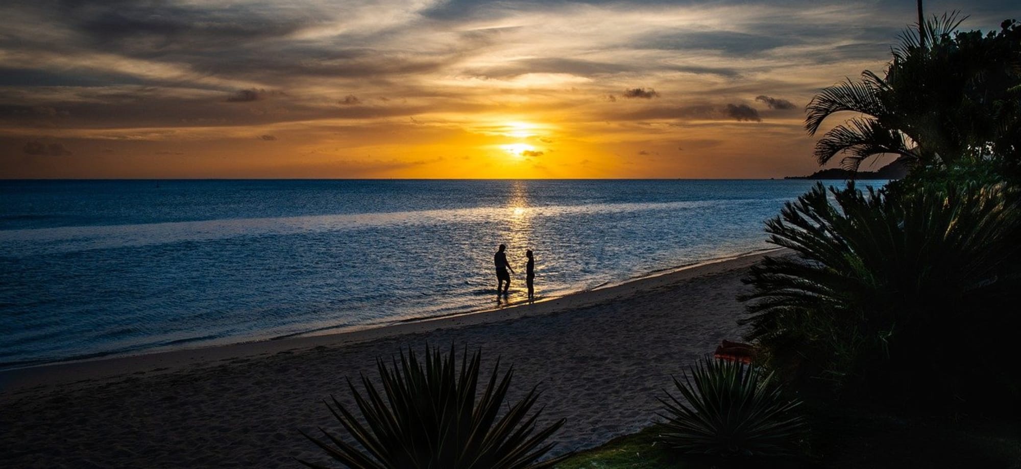 A couple are walking along a beach at night when the sun is setting. 