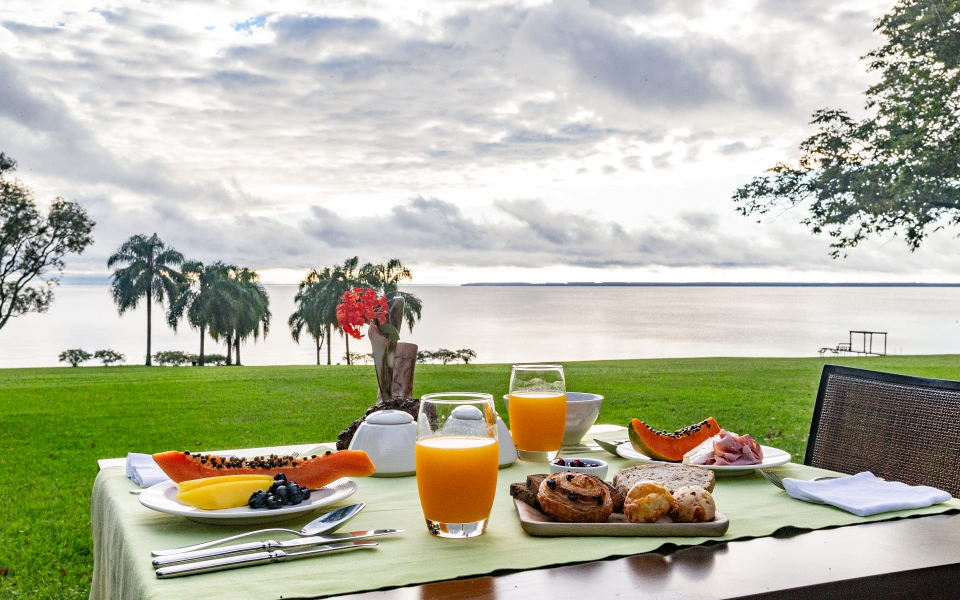 A two-person table is set up on the lawn, overlooking the water and palm trees, with breakfast, pastries and orange juice.