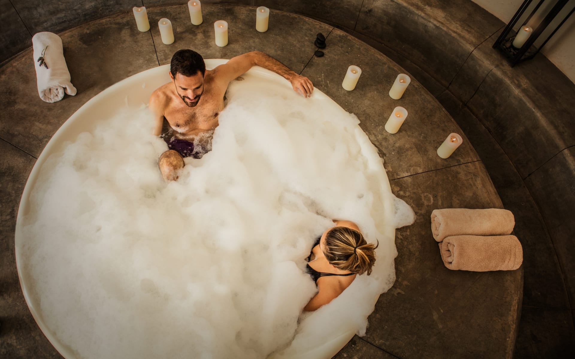 A young couple are enjoying a foaming plunge in Canvas Wine Lodge's jacuzzi, which is surrounded by towels and candles. 