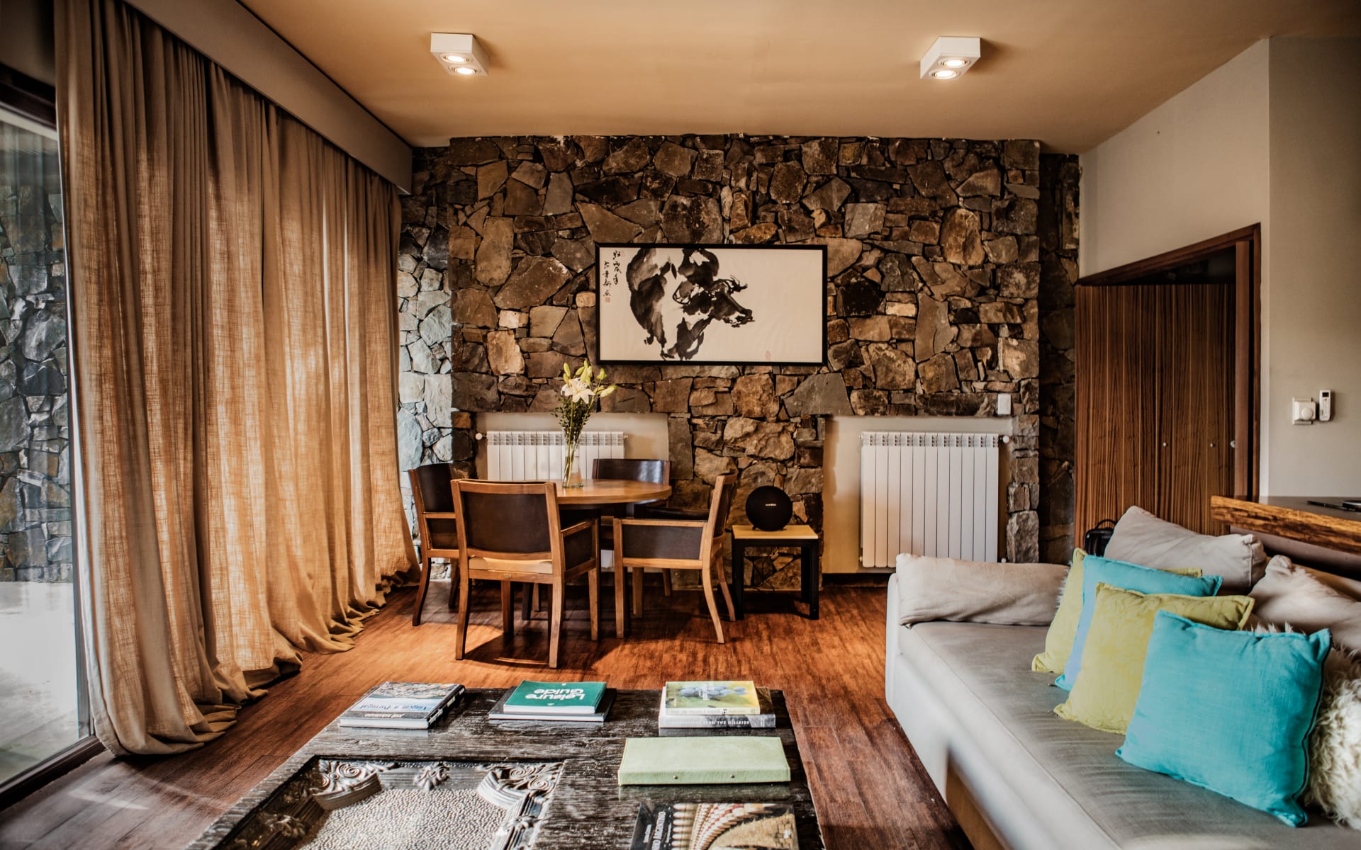 Canvas Wine Lodge Villas have a lounge with floor-to-ceiling windows, a stone wall featuring contemporary artwork and a dining table and sofa.