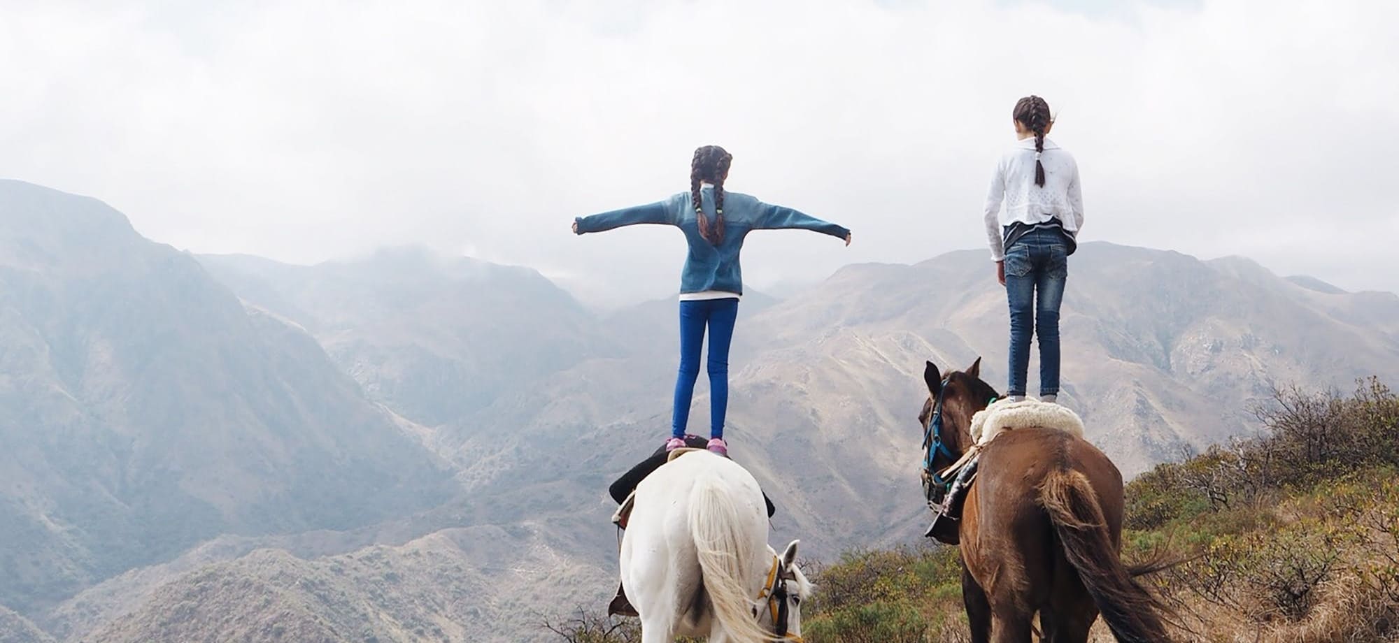Two young girls are standing on horses' backs overlooking mountains. 