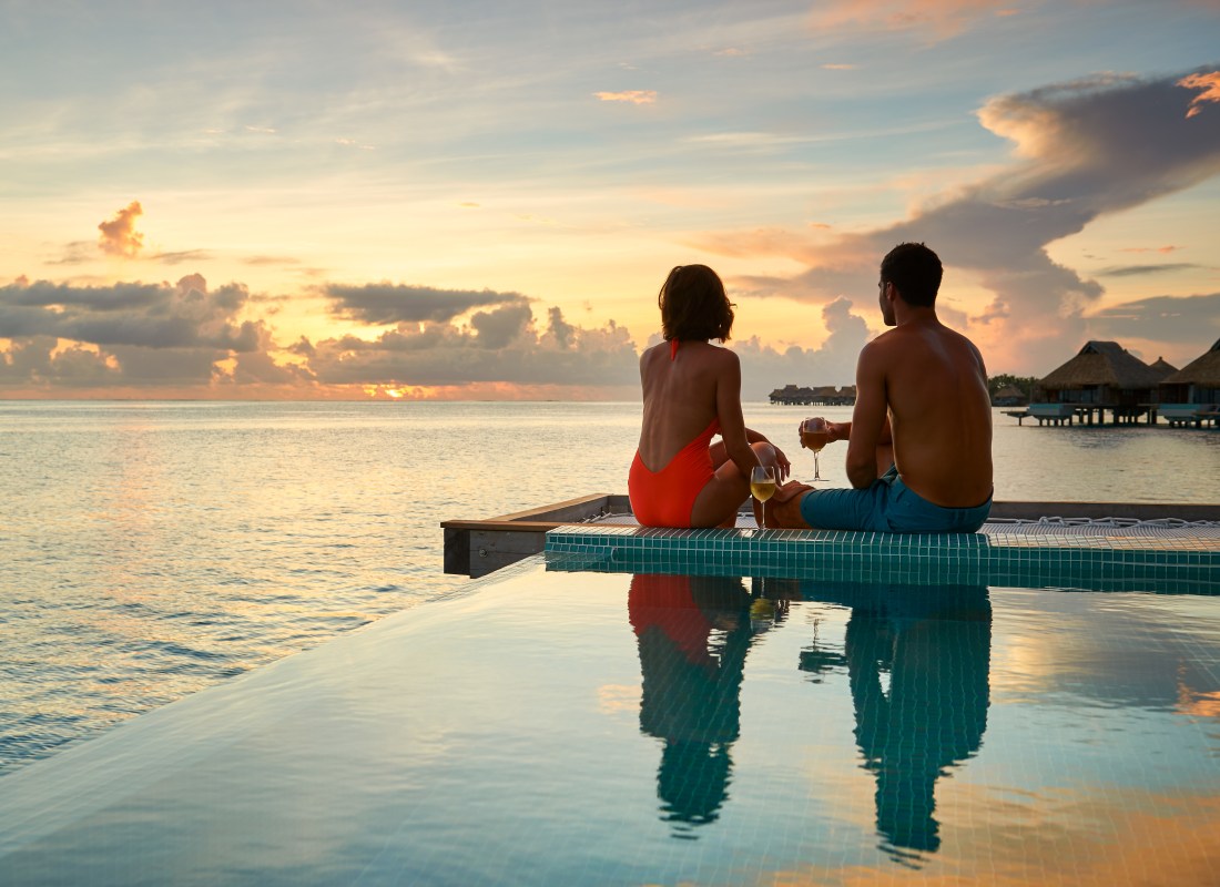 A couple are sitting on the edge of an infinity pool watching the sun set. 