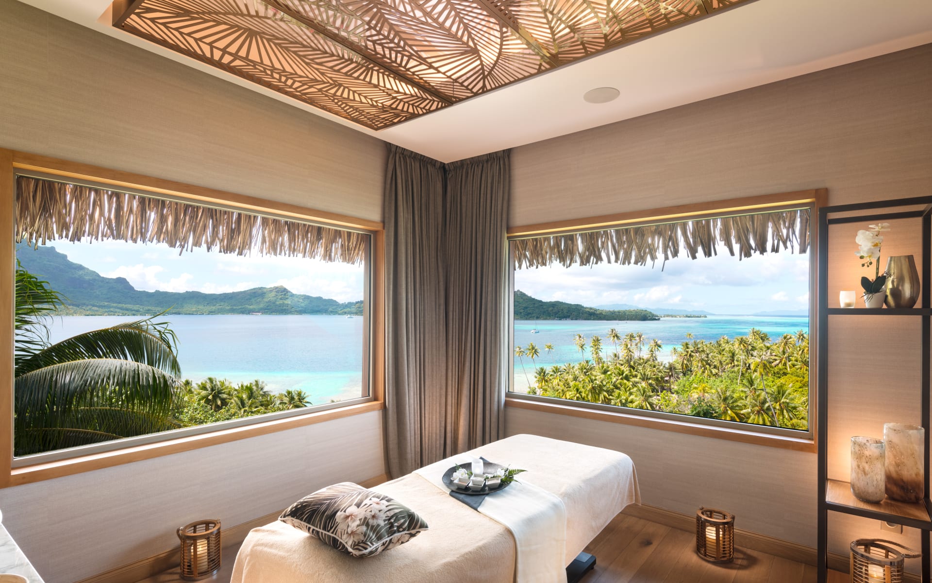 The spa room has a single bed with a pillow, surrounded by candles and two windows with ocean views. 