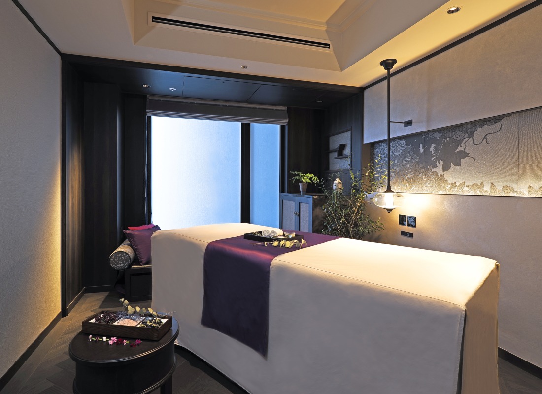 There is a single spa table in a box room with purple cushions, flowers and hot towels. 