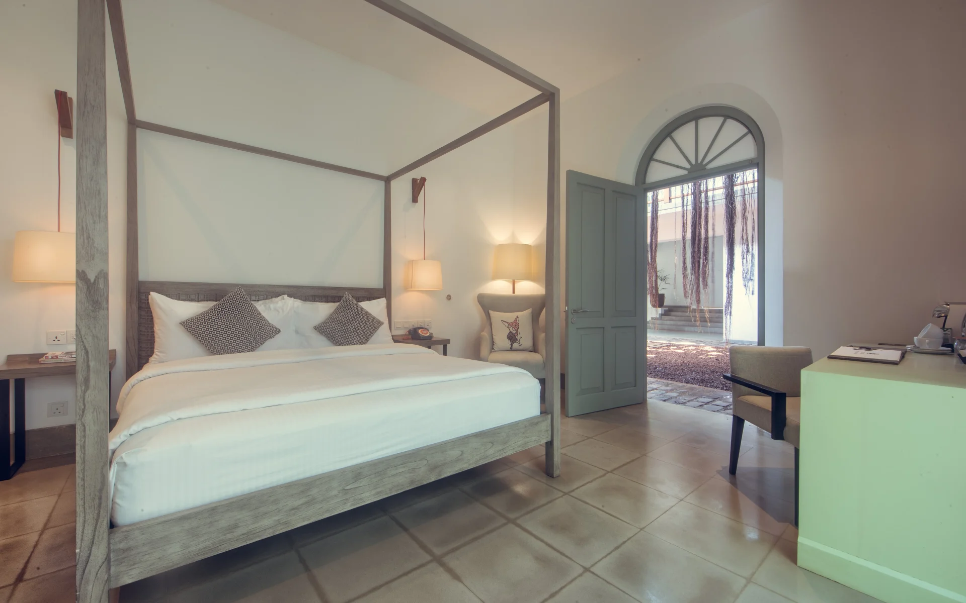 One of the bedrooms has glossy, tiled flooring and a large, four-poster bed dressed in white linen bedding. The door leads directly outside.