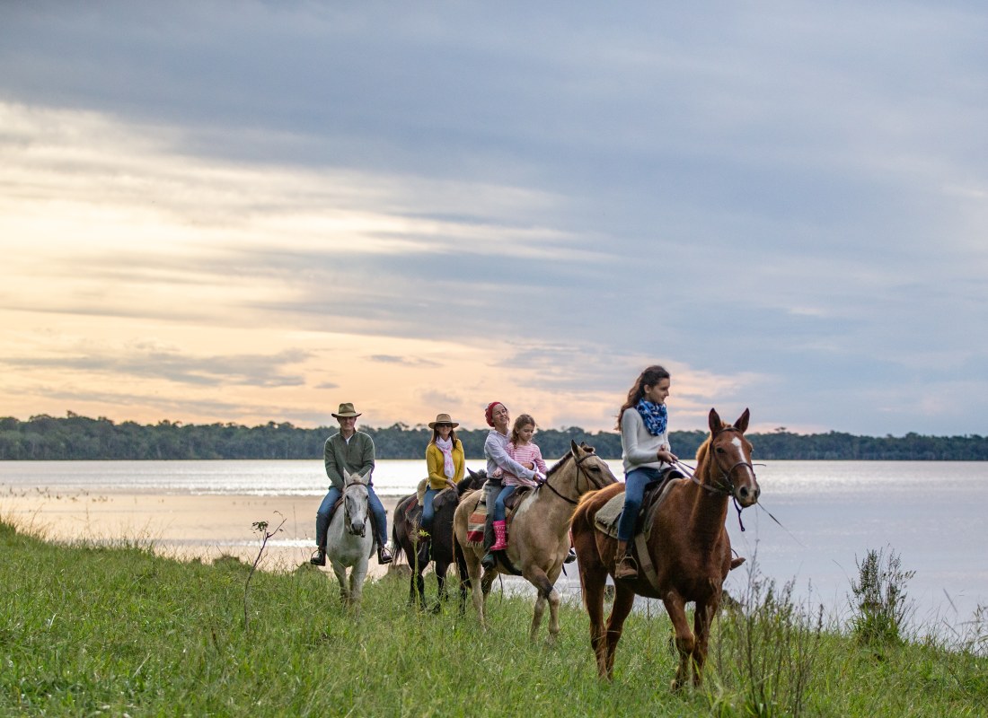 Four adults and a child are horseriding next to the water as the sun sets. 