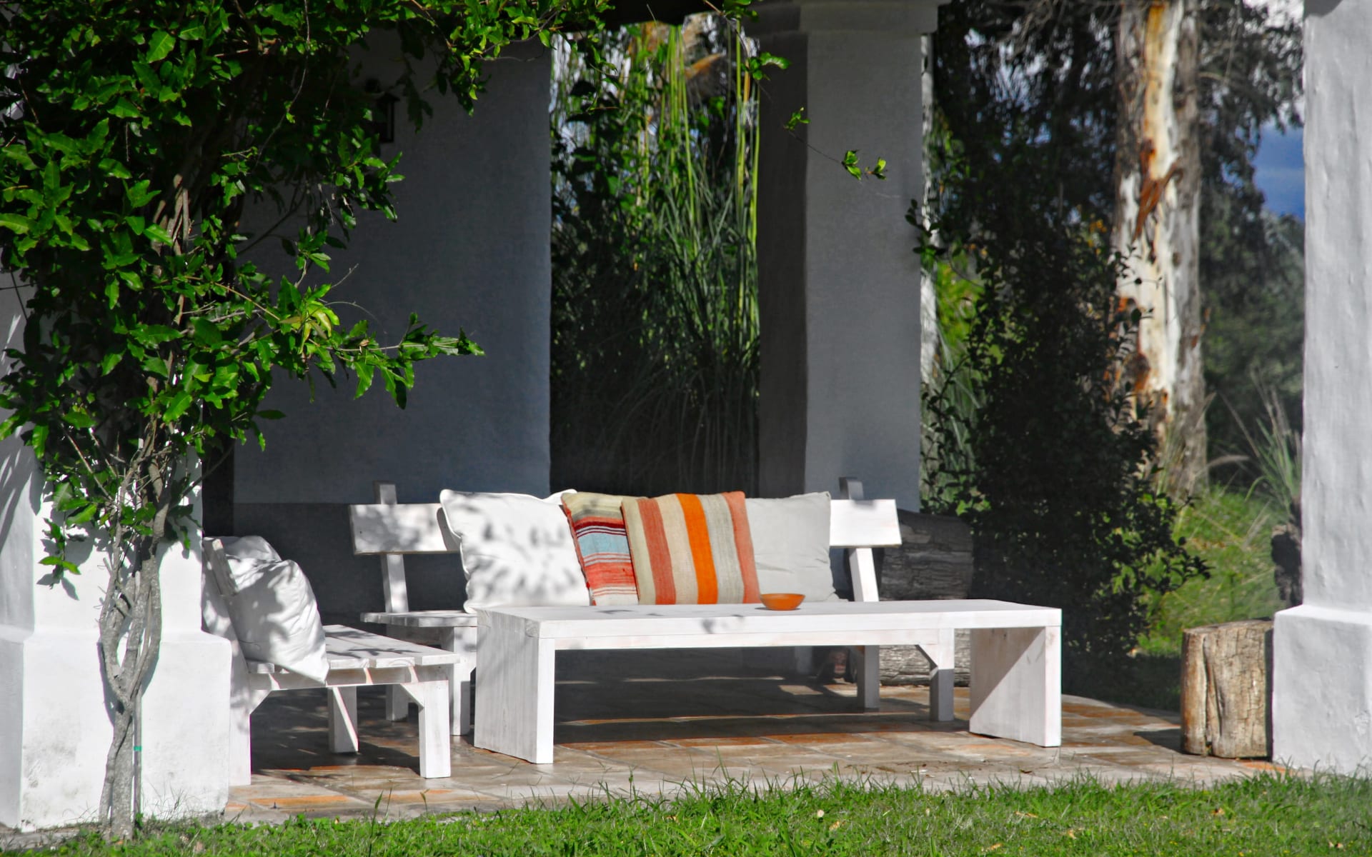 Trees surround a white bench with orange stripped cushions and a white table overlooking a green lawn. 