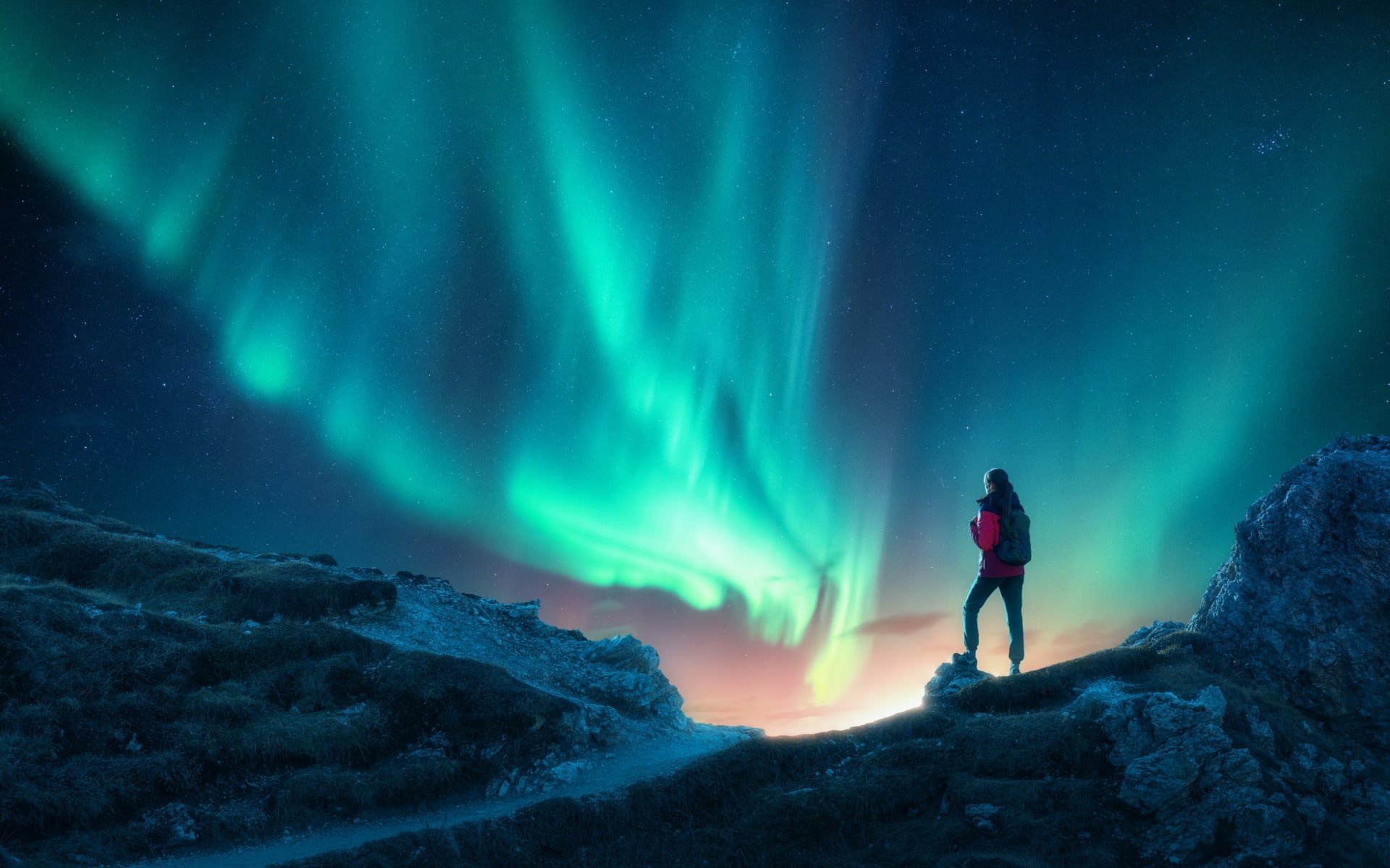 A woman stands on a rocky high-point, watching as the sky turns vibrant blues and greens on a Northern Lights display.