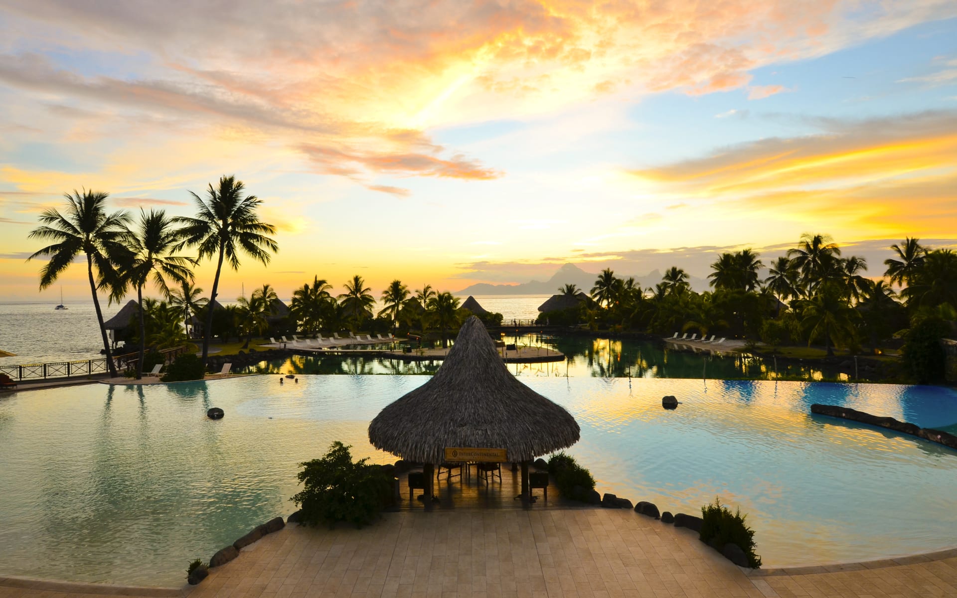 The sun is setting on the swimming pool, backed by the ocean and palm trees. 